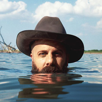 Marty Bush rising up out of a lake while wearing a cowboy hat. He is submerged up to his beard and facing the viewer.
