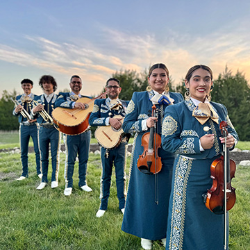 Mariachi Estrella members wearing traditional clothing are standing in a line and smiling at the viewer
