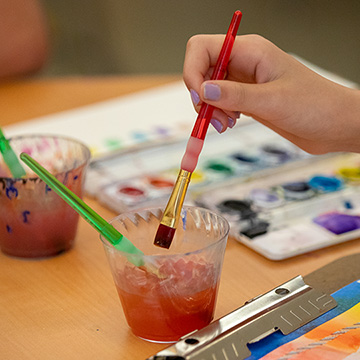 A child's hand dipping a paint brush in a cup of water