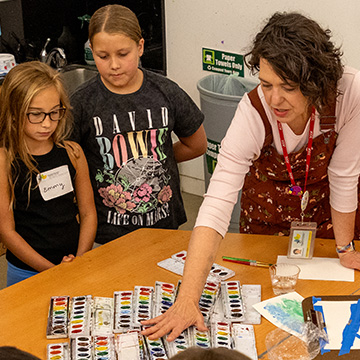 a group of students stand around a table while an instructor point to watercolor laid out on the table