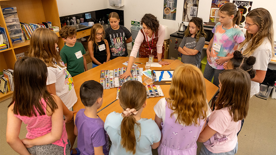 a group of students stand around a table while an instructor point to watercolor laid out on the table