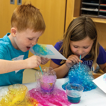 two young children blow colorful bubbles during an art class