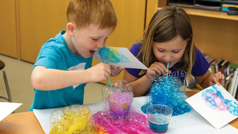 two young children blow colorful bubbles during an art class