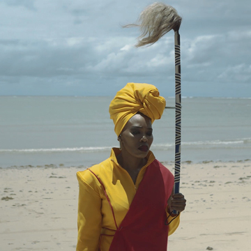 A woman in a yellow dress and head wrap stands on the beach holding a striped rod with a tuft of hair at the top. Her face in in shadow and she looks solemn with her eyes closed. She is facing away from the water.